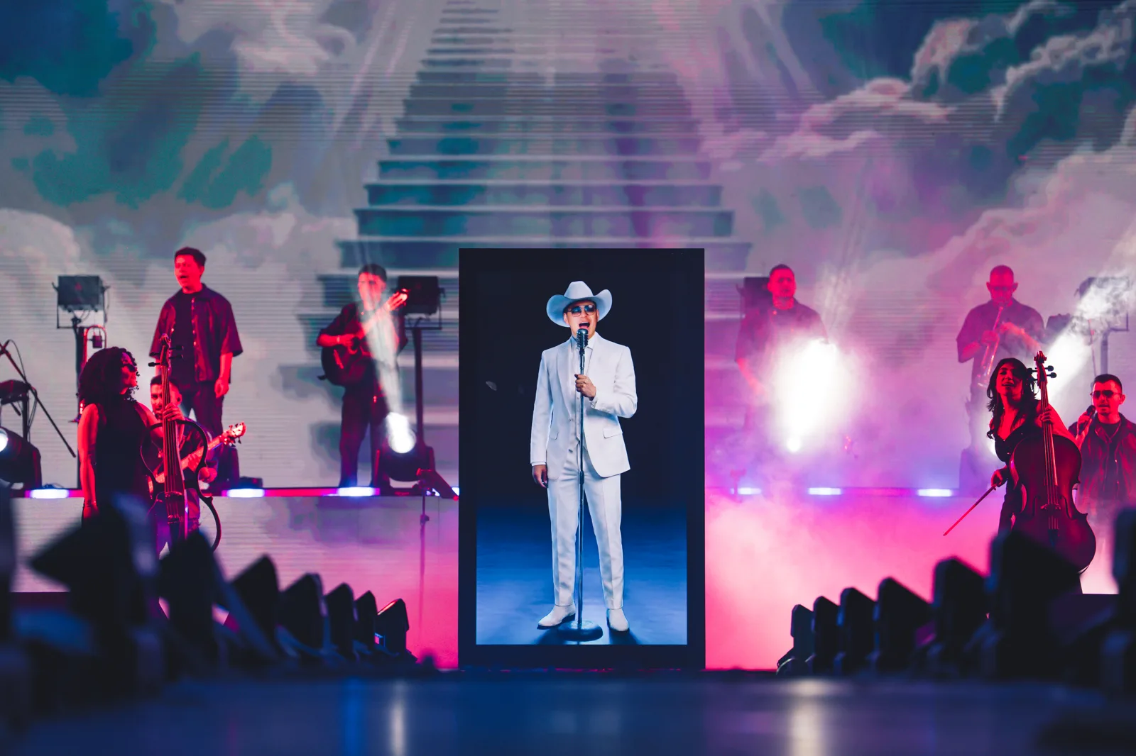 El avatar con IA de Yeison Jiménez, vestido de blanco, dentro del marco central mientras su banda toca en vivo bajo la escalinata proyectada, en el concierto homenaje de El Campín
